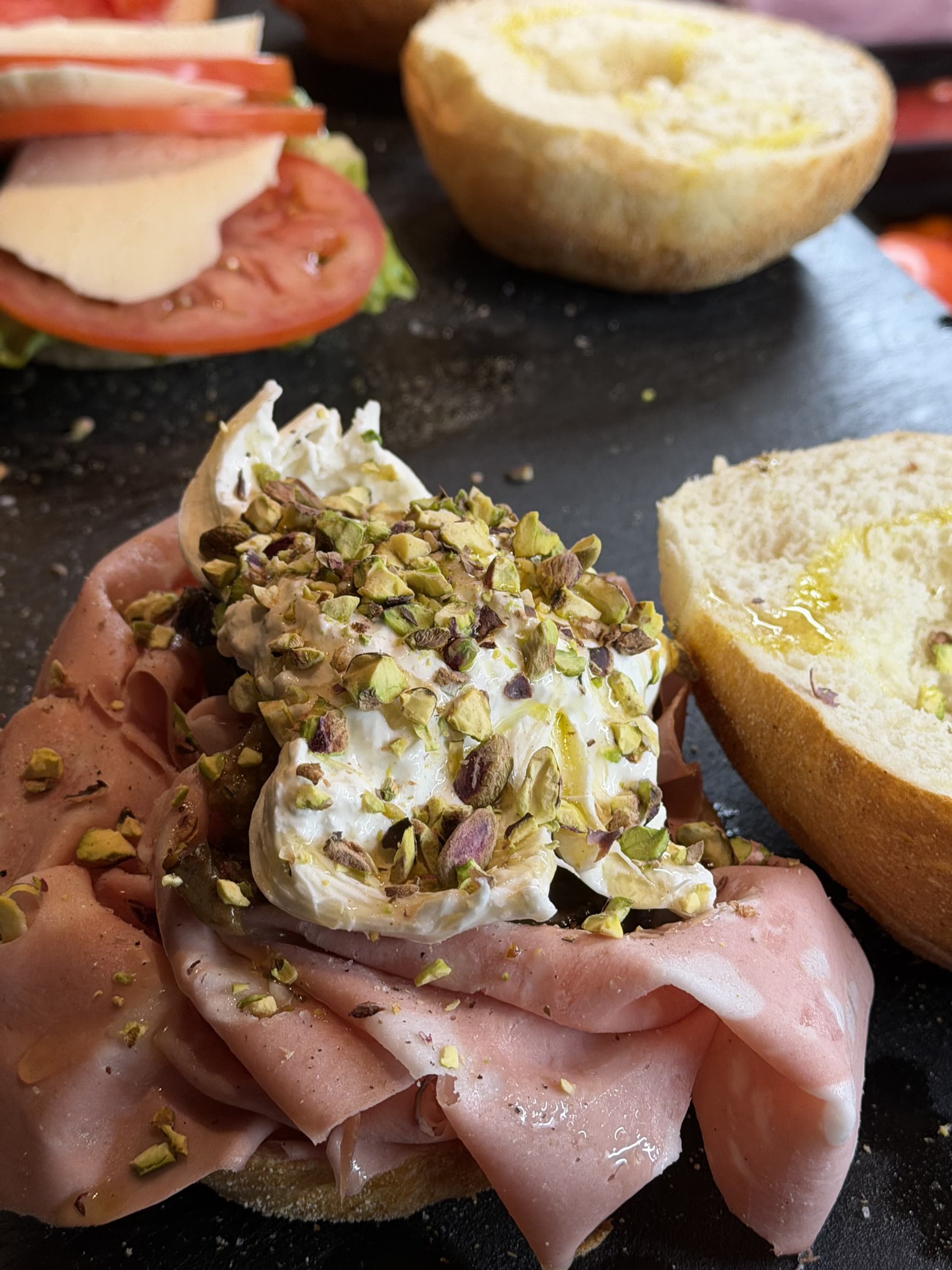 Burrata toppas med hackad pistage och olja ovanpå skivad skinka, bakom disken.