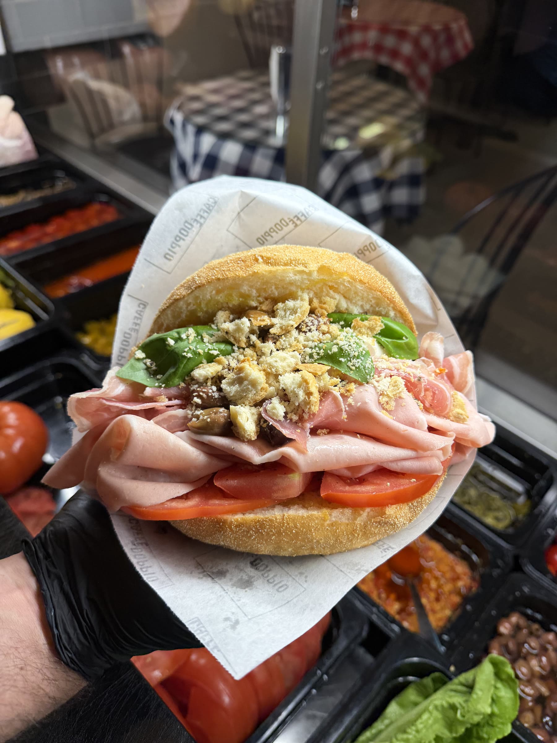 En färdig smörgås med mortadella, prosciutto, tomat, basilika och taralli i DOPPIO-märkt papper, hållen över disken.