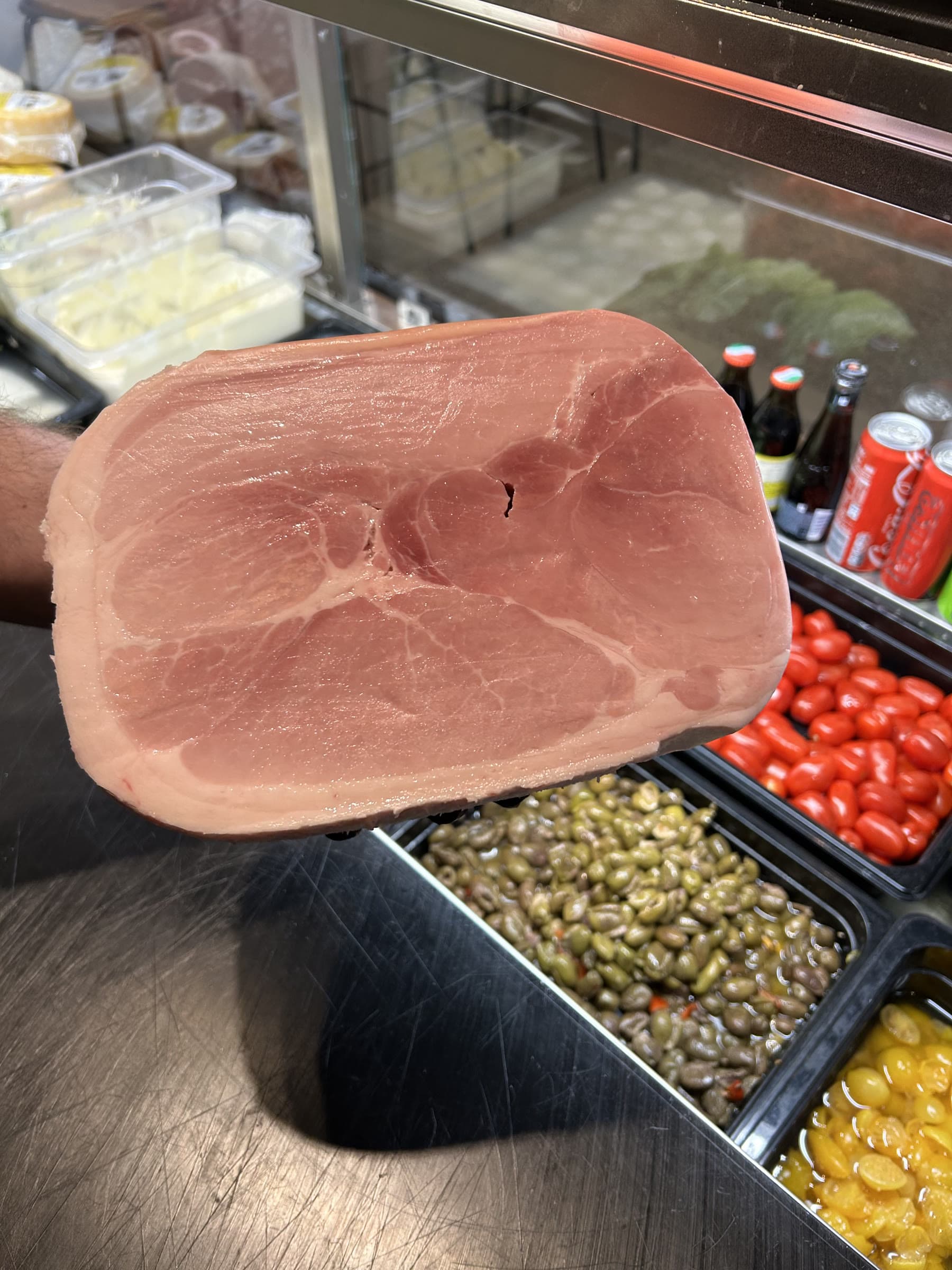 Ett helt kokt skinklår hålls upp framför oliver och körsbärstomater i disken.
